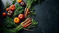 Fresh organic vegetables including heirloom tomatoes, rainbow carrots, and leafy kale arranged on dark slate surface for healthy cooking and farm-to-table cuisine.