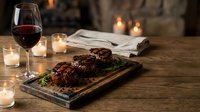 Grilled steak dinner with red wine and candles on rustic wooden table creating cozy romantic dining atmosphere by warm fireplace.