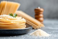 Fresh spaghetti pasta on black plate with grated parmesan cheese and raw noodles, wooden pepper mill in background on gray concrete surface.