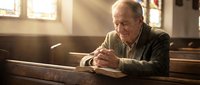 Elderly Caucasian man praying peacefully in wooden church pew with warm sunlight streaming through stained glass windows creating spiritual atmosphere.