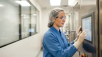Female scientist in blue lab coat and hair net operating touchscreen control panel in modern sterile laboratory facility for pharmaceutical research.
