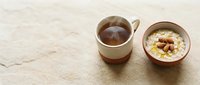 Hot tea in ceramic cup and bowl of oatmeal with nuts on beige textured surface, overhead flat lay composition for breakfast, food styling, and culinary content.