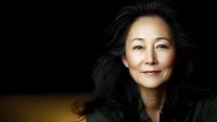 Mature Asian woman with long black hair smiling warmly against dark background, natural makeup and gentle expression portrays confidence and wisdom. Professional portrait lighting.
