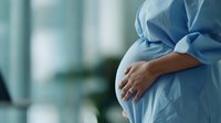 Pregnant woman in light blue dress gently holding her belly against blurred hospital background, side view close-up showing tender maternal moment and expectant care.