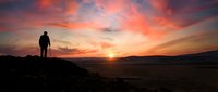 Silhouette of man standing on mountain peak watching dramatic sunset with orange and pink clouds over desert landscape for inspiration and achievement concepts.