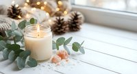 Cozy winter spa scene with burning candle, eucalyptus leaves, pink salt crystals and pine cones on white wooden surface with warm bokeh lights.