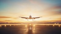Airplane landing at sunset on illuminated runway with dramatic orange sky and city silhouette in background, front view perspective of descending aircraft.