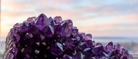 Stunning amethyst crystal cluster with deep purple gemstone points against a soft pastel sunset sky background, showcasing natural mineral beauty.