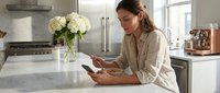 Young woman with medium skin tone, casual attire, reading magazine at kitchen counter with white flowers and modern appliances, lifestyle home interior.