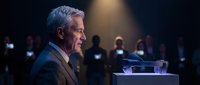 Senior male executive with gray hair and light skin tone speaking at podium during corporate conference with audience holding mobile devices in background.
