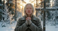 Young blonde woman in warm winter coat praying peacefully in snowy forest during golden hour sunset with magical bokeh lights creating serene spiritual atmosphere.