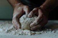 African American hands kneading raw dough on floured surface, close-up view showing traditional bread making process and flour-covered fingers at work.