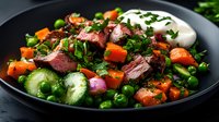 Juicy grilled steak slices with roasted vegetables, green peas, cucumber, and sour cream on black plate garnished with fresh herbs, close up food photography.
