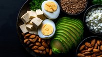 Healthy ketogenic diet ingredients with sliced avocado, boiled eggs, nuts, seeds and cottage cheese arranged on dark wooden board for low carb meal prep.