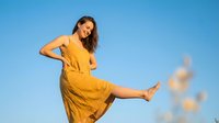 Young woman in yellow dress dancing joyfully against clear blue sky during golden hour sunset. Carefree lifestyle and happiness concept for wellness content.