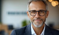Confident mature Caucasian businessman with gray hair and glasses smiling at camera. Professional portrait with soft bokeh background in modern office setting.
