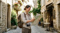 Young Caucasian male tourist with backpack reading map on cobblestone street in historic Mediterranean old town with stone buildings and outdoor cafe.