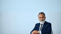 Middle-aged man with gray beard and hair, South Asian ethnicity, wearing dark navy business suit and light blue tie, seated with hands clasped, against plain light blue background for corporate and professional use.