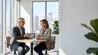 Two professionals in business casual attire discuss work using a tablet at a small table in a modern office with floor-to-ceiling windows overlooking a city skyline and potted plants nearby.