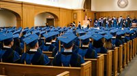 Graduation ceremony with students in blue caps and gowns seated in wooden auditorium preparing for commencement celebration and academic achievement.