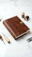 Handmade leather journal with brass clasp surrounded by bookbinding tools and thread spools on marble surface for crafting and writing projects.