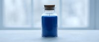 Blue liquid in glass bottle with cork stopper on white surface against blurred window background for laboratory research and chemical experiments.