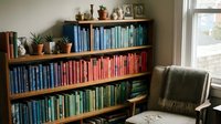Cozy home library with color-coordinated books arranged on wooden shelves, featuring small potted plants and comfortable reading chair by window.