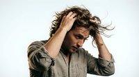 Young man with curly dark hair and olive complexion wearing olive linen shirt, hand in hair, looking downward with contemplative expression against white background.