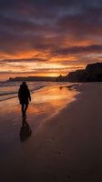 Solitary figure in wetsuit walking along sandy beach at dramatic golden sunset with rocky cliffs, reflecting light on wet sand, ideal for travel and lifestyle content.