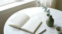Minimalist workspace with open notebook, wooden pen and eucalyptus branch in ceramic vase on white marble table near window for journaling and writing.