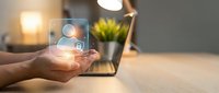 Hand holding glowing digital security shield icon with lock symbol over modern workspace with laptop and warm lighting for cybersecurity concepts.