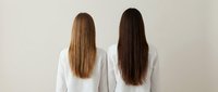 Two women with long straight hair standing back to camera wearing white shirts against neutral background for hair care and beauty concepts.