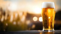 Cold glass of light beer with foam head and bubbles against blurred sunset background, golden hour lighting creates atmospheric mood for beverage photography.