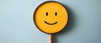 Yellow round smiley face on wooden stick against light blue wall background, representing happiness, positivity and cheerful emotions in minimalist style.