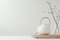 White ceramic teapot on wooden tray with delicate cherry blossom branch in glass vase against neutral wall, creating minimal zen atmosphere for wellness concept.