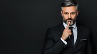Confident mature businessman with gray hair and beard wearing black suit and tie thoughtfully touching face against dark background. Professional portrait.