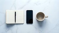 Minimalist workspace flat lay with open notebook, black pen, smartphone and empty coffee cup on white marble surface for productivity and planning concepts.