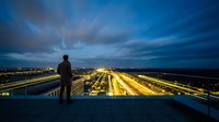 Silhouette of man standing on rooftop overlooking illuminated highways and city lights at dusk, urban landscape with dramatic sky for business, success, and vision concepts.