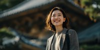 Young Asian businesswoman in gray blazer smiling confidently outdoors with traditional architecture background for corporate and professional concepts.