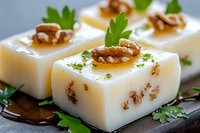 Elegant appetizer cubes of creamy white cheese garnished with walnuts, fresh herbs and honey drizzle on dark serving board, macro food photography.