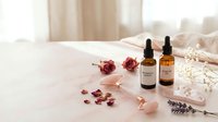 Skincare essential oils and natural ingredients arranged on white fabric with dried flowers, rose quartz roller, and lavender for wellness and beauty routines.