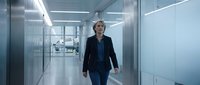 Middle-aged Caucasian male professional in dark business suit walking through modern corporate office corridor with glass partitions and fluorescent lighting.