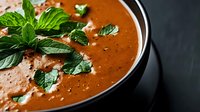 Creamy tomato soup garnished with fresh mint leaves and croutons in a black bowl, ready for lunch or dinner serving.