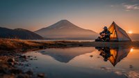 Solo camper sits by tent near mountain lake at golden hour sunrise with perfect water reflections creating serene wilderness camping scene.