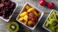 Colorful dried fruits and fresh kiwi slices arranged in white ceramic bowls on rustic wooden background, healthy snacks and natural food styling.