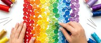 Hands arranging colorful buttons in a rainbow gradient pattern on white surface, surrounded by thread spools and needles for sewing.