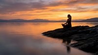 Woman sitting on rocky outcrop at sunset over calm water, silhouetted against golden sky with mountains in distance. Peaceful meditation and contemplation scene.