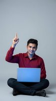 Young Indian businessman in red shirt pointing up with laptop computer sitting cross-legged on floor showing eureka moment and creative thinking.