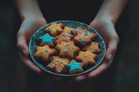 Female hands holding plate with homemade star-shaped sugar cookies decorated with blue icing and colorful sprinkles against dark moody background.