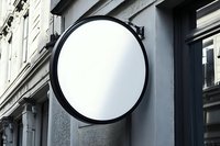 Round blank white signboard with black frame mounted on gray building facade, mockup for store or restaurant branding in urban setting.